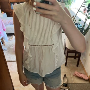 Vit blus med volangdetaljer - Supersöt vit blus i bomull med volanger längs sidorna och axlarna. Framsidan har smala veck och en liten spetskantad detalj under bysten. Ärmlös modell med lätt v-ringning i ryggen. Perfekt för en fräsch och somrig look!