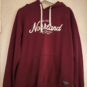 Vinröd hoodie från Norrland - Snygg vinröd hoodie från The Great Norrland med vit text på bröstet och vita snören i huvan. Klassisk känguruficka framtill och mjukt material som passar perfekt för kyliga dagar. Enkel och stilren design med avslappnad passform.