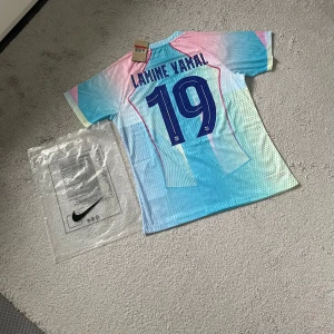 FC Barcelona Lamine Yamal Nike tröja - Säljer en FC Barcelona fotbollströja med Lamine Yamal och nummer 19 på ryggen. Tröjan har en unik design i ljusblått, rosa och inslag av vitt, med klubbmärke, Nike-logga och Spotify som sponsor. Tillverkad i lätt och ventilerande material, perfekt för match eller träning.