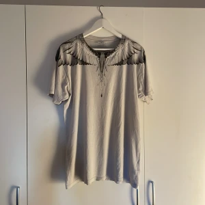 Vit t-shirt med vingar från Marcelo Burlon - Snygg vit t-shirt från Marcelo Burlon med ikoniskt vingtryck i svart och grått över axlar och rygg. Klassisk passform, rund hals och tillverkad i 100% bomull. Perfekt statement-plagg för dig som gillar streetwear vibes.