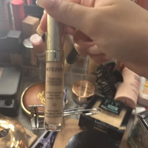 By Terry concealer i ljus nyans - By Terry concealer med guldig kork och transparent tub som visar en ljus beige krämig formula. Perfekt för att täcka mörka ringar och ge en fräsch look. Enkel att applicera med applikatorn i locket.