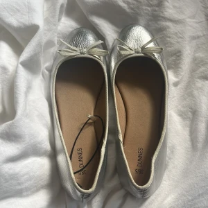 Silvriga ballerinaskor med rosett från Dunnes - Snygga ballerinaskor i metallic-silver från Dunnes med rund tå och dekorativ rosett framtill. Dem är storlek 38/5 men sitter mer som 37 skulle jag säga.