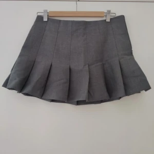 Grå plisserad kortkjol - Snygg grå kortkjol med breda plisserade veck. Kjolen har en klassisk och stilren look som passar perfekt till sneakers eller boots. Enkel att matcha med både stickade tröjor och t-shirts för en trendig vibe.