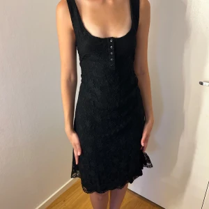 Svart spetsklänning med knappar - Supersnygg svart kortklänning i spets med djup urringning och dekorativa knappar framtill. 