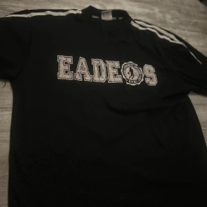 Svart tröja Eades med vita ränder - Svart tröja från Eades Wear med vita ränder längs axlarna och tryckt text 'EADEOS' på bröstet. Snygg sportig look med nummer 32 tryckt på ryggen. Perfekt för dig som gillar college-stil och streetwear.