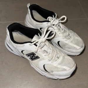 Vita New Balance 530 sneakers - Snygga New Balance 530 sneakers i vitt med svarta detaljer. Skorna har meshpaneler för extra andningsförmåga, klassisk snörning och en chunky sula med ABZORB-dämpning. Perfekta för dig som gillar sportig och trendig stil.