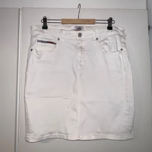 Vit jeanskjol från Tommy Jeans - Vit jeanskjol från Tommy Jeans med klassisk femficksdesign och snygga detaljer i rött och blått på fickorna. Kjolen har en liten slits bak och logopatch på linningen. Perfekt för en fräsch och stilren look. Använd fåtal gånger. Passar M-XL skulle jag säga. Strechig❤️