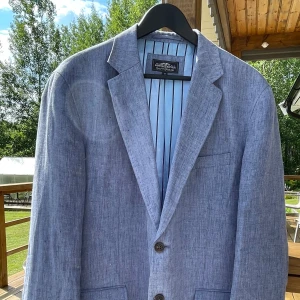 Blå linnekavaj Hampton Republic 56 - Snygg blå kavaj från Hampton Republic i storlek 56. Tillverkad i linne med klassisk passform, två knappar framtill och flera fickor. Insidan har ett stilrent blått foder med svarta ränder. Perfekt för en fräsch och avslappnad look.