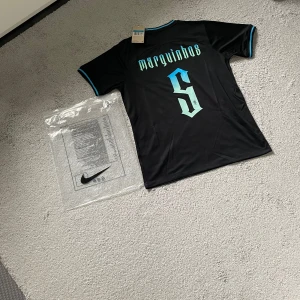 Svart Brasilien Marquinhos Nike tröja - Snygg svart Brasilien fotbollströja från Nike med Marquinhos och nummer 5 på ryggen. Tröjan har turkosa detaljer vid krage och ärmslut, samt tryckt klubbmärke och Nike-logga på bröstet. Tillverkad i lätt och ventilerande material, perfekt för match eller träning.