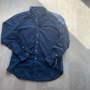 Marinblå skjorta från Ralph Lauren - Klassisk marinblå skjorta från Ralph Lauren med button-down krage och vit knappstängning. Skjortan har lång ärm och den ikoniska röda broderade loggan på bröstet. Perfekt för en stilren och avslappnad look. Hål i armen 