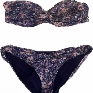 Blommig bandeau bikini i lila och rosa - Supersöt bikini med bandeau-topp och matchande trosa i lila och rosa blommigt mönster. Twistad detalj framtill på både överdel och underdel. Perfekt för stranden eller poolen, ger en trendig och feminin look.