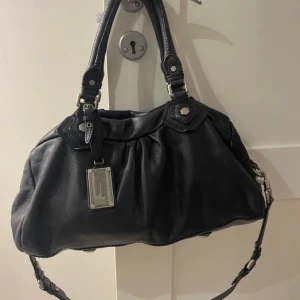 Classic Q Groovee satchel från Marc by Marc Jacobs  - Väskan på bilden är en svart läderväska, specifikt en Classic Q Groovee satchel från Marc by Marc Jacobs med silverdetaljer. Passar perfekt som både en vardagsväska men även en väska man kan resa med som handbagage 🥰 Perfekt till allt 🩵 Den är svår att hitta eftersom den tyvärr inte säljs längre. Defekter finns med på bilderna