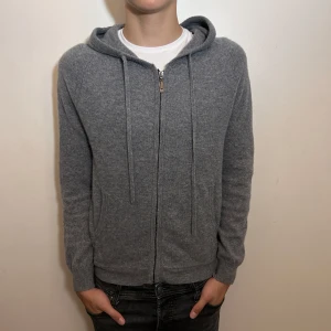 Grå hoodie i kashmir med dragkedja - Mysig grå hoodie i kashmir med hel dragkedja framtill och snörning i huvan. Tröjan har två fickor och ribbade muddar vid ärmslut och nederkant. Perfekt för dig som gillar en clean och enkel stil med lyxig känsla. 100% Kashmir