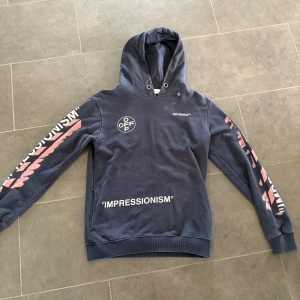 Off-White blå hoodie med tryck - Säljer en blå hoodie från Off-White med huva och feta tryck i vitt och rosa på både ärmar, bröst och rygg. Stora grafiska detaljer och texten 'IMPRESSIONISM' ger en riktigt street vibe. Perfekt för dig som vill sticka ut med en ikonisk look.