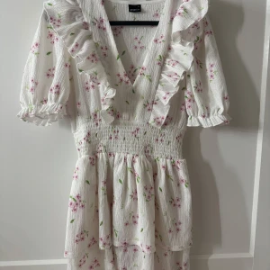 Vit blommig volangklänning Gina Tricot - Supersöt vit kortklänning från Gina Tricot med rosa blommor och gröna blad. Klänningen har v-ringning, volangdetaljer längs urringningen och ärmarna, samt smock i midjan. Perfekt för sommarens alla tillfällen!