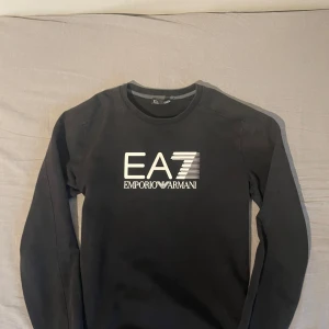 Svart EA7 Emporio Armani tröja - Svart långärmad tröja från EA7 Emporio Armani med vit logga framtill. Klassisk rund halsringning och ribbade muddar vid ärmslut och nederkant. Mjuk och bekväm, perfekt för en stilren look.