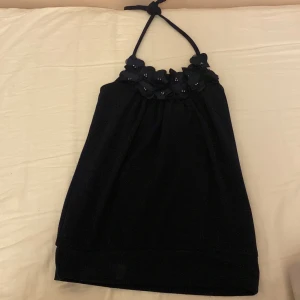 Svart halterneck topp med blommor - Supersöt svart halterneck topp med dekorativa blommor och pärldetaljer längs halsringningen. Toppen är ärmlös och har knyt i nacken för en snygg passform. Perfekt för dig som vill sticka ut med en unik och feminin look. XS/XSS