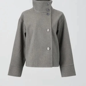 Grå kappa med hög krage - Snygg grå kappa med snygg passform och breda ärmar. Jackan har en hög krage med flera knappar och kan bäras både uppknäppt och stängd. Perfekt för dig som gillar minimalistisk stil och vill ha något som passar in i trender!