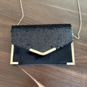 Svart kuvertväska med paljetter och kedja - Snygg svart kuvertväska med glittrig paljettflik och guldfärgade metalldetaljer i hörnen. Väskan har en elegant kedjerem i guld och stilren, rektangulär form. 