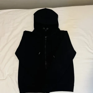 Svart hoodie med dragkedja och huva - En stilren svart hoodie med dragkedja framtill och justerbar huva med snörning. Tröjan har långa ärmar och ribbade muddar vid ärmslut och nederkant. Perfekt för en chill och avslappnad look.