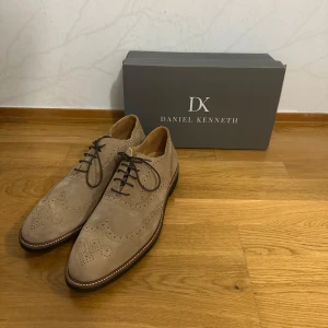 Beige mockaskor från Daniel Kenneth - Snygga beige mockaskor från Daniel Kenneth med klassiskt broguemönster och rund tå. Skorna har mörka snören och en platt sula med kontrastsöm runt om. Perfekta för dig som vill ha en dressad men ändå chill look.