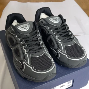 Dior B30 - Säljer ett par svarta Dior B30 sneakers med vita detaljer och reflekterande inslag som ger skorna en exklusiv och modern look. Modellen har klassisk snörning, rund tå och en chunky platt sula som både ger komfort och stil. Extra snören ingår. Hör av dig vid frågor och funderingar!