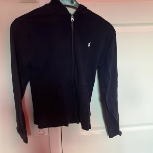 Svart hoodiejacka från Ralph Lauren XL - Mörkblå hoodiejacka från Polo Ralph Lauren i storlek XS. Jackan har dragkedja framtill, huva och två fickor. Klassisk vit logga broderad på bröstet. Tillverkad i mjuk bomull, perfekt för en chill och stilren look.