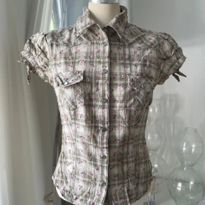 Rutig blommig skjorta med knappar - Kortärmad skjorta i beige och gröna toner med rutigt mönster och små rosa blommor. Skjortan har tryckknappar framtill, två bröstfickor och knytband på ärmarna. Figurnära passform och klassisk krage. Perfekt för dig som gillar en söt och avslappnad stil.