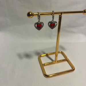 Hjärtformade örhängen med röda stenar - Snygga örhängen i silverfärg med hjärtformade hängen. Hängena har en röd glittrande sten i mitten och är omgivna av små klara stenar som ger extra bling. Perfekta för dig som vill sticka ut med en romantisk och trendig look.