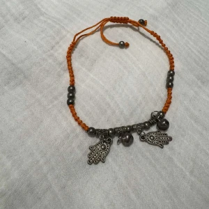 Orange ankellänk med hamsa-berlocker - Unikt armband med orange flätad tråd, dekorerat med silvriga pärlor och tre detaljerade hamsa-händer som berlocker. Justerbar knytning för perfekt passform. Perfekt accessoar för dig som gillar boho-stil och symboliska smycken.