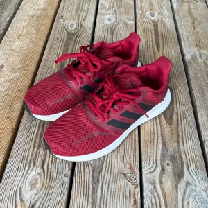 Röda Adidas sneakers - Snygga röda Adidas sneakers med svart detalj och klassiska tre ränder på sidan. Ovandelen är i ventilerande mesh och skorna har snörning samt vit platt sula. Perfekta för dig som gillar sportig stil och vill ha bekväma skor till vardagen.