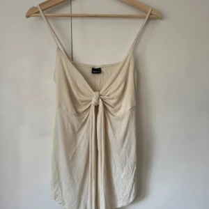 Beige linnetopp med knyt fram - Supersöt beige linnetopp från Monki med smala axelband och snygg knytning framtill. Toppen har en avslappnad passform och är tillverkad i mjuk bomull, perfekt för varma dagar. Enkel men ändå trendig design som passar till jeans eller kjol.