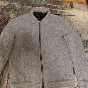 Grå mönstrad overshirt med dragkedja - Snygg grå overshirt med diskret fiskbensmönster och dragkedja framtill. Jackan har klassisk krage, lång ärm och en bröstficka. Perfekt för lager-på-lager och passar till jeans eller linnebyxor storlek. Storlek S