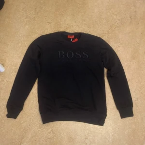 Hugo boss tröja  - Svart sweatshirt från Hugo Boss med stor BOSS-logga broderad på bröstet. Tröjan har rund halsringning, långa ärmar och ribbade muddar. Perfekt för dig som gillar stilren och klassisk streetwear med premiumkänsla.