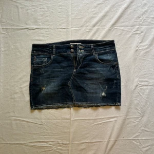Mörkblå jeanskjol med slitningar - Midjemått rakt över: 40 cm, längd: 33 cm Snygg mörkblå jeanskjol med slitna detaljer och rå kant nertill. Klassisk femficksmodell med dubbla knappar framtill och broderade bakfickor. Perfekt för en avslappnad och trendig look.