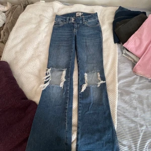 Blå bootcut jeans med slitningar 157 - Bootcut jeans från 157 i blå tvätt med stora slitningar och hål över knäna. Klassisk femficksmodell, normal passform och midja. Jeansen är i bomull och har en cool, avslappnad vibe. Perfekta för dig som gillar en edgy streetstil.