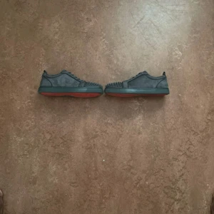 Christian Louboutin grå mocka sneakers - Säljer ett par grå sneakers från Christian Louboutin i mocka med röda sulor och coola nitar framtill. Skorna har snörning och rund tå, samt den klassiska Louboutin-loggan på insidan. Perfekta för dig som vill sticka ut med exklusiva detaljer.
