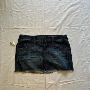 Mörk jeanskjol med rå kant - Midjemått rakt över: 40 cm, längd: 30 cm Snygg mörkblå jeanskjol i minilängd med rå, fransig kant nertill. Klassisk femficksmodell med bälteshällor och knappstängning framtill. Perfekt till en enkel topp eller hoodie för en chill look.