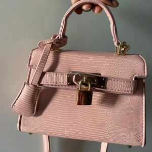 Liten ljusrosa Kelly väska - Säljer en superfin rosa Kelly väska med krokodilmönster och guldfärgade detaljer. Väskan har både handtag och justerbar axelrem, samt snyggt lås framtill. Helt oanvänd, pris kan diskuteras vid snabb affär 🩷🩷