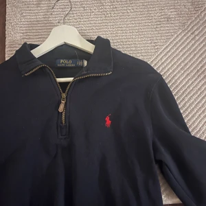 Marinblå half zip från Polo Ralph Lauren - Snygg marinblå långärmad tröja från Polo Ralph Lauren med klassisk krage och half zip-dragkedja. Tröjan har det ikoniska röda broderade logot på bröstet och är gjord i mjuk bomull. Perfekt för en stilren och avslappnad look.