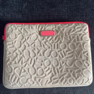 Datorfodral Marc by Marc Jacobs - Säljer en beige datorväska från Marc by Marc Jacobs med coolt 3D-bokstavsmönster och rosa detaljer. Fläckarna går bort om man tvättar