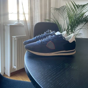 Blå sneakers från Philippe Model - Snygga blå sneakers från Philippe Model med vita detaljer och röd kant runt loggan på sidan. Ovandelen är i mocka och textil, med vit häl och klassisk snörning. Skorna har en platt sula och rund tå, perfekt för en stilren och sportig look.