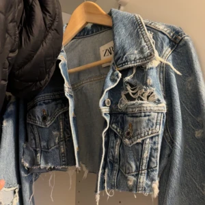 Croppad jeansjacka från Zara - Säljer en croppad ljusblå jeansjacka från Zara med slitna detaljer och råa kanter. Jackan har klassisk krage, bröstfickor med knappar och lång ärm. Har använt den ungefär 3-4 ggr bara 