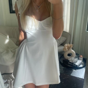 Vit kort klänning - Supersöt vit kort klänning med markerad byst och lätt utsvängd kjol. Klänningen har breda axelband och en figurnära passform upptill som ger en feminin siluett. Perfekt för sommarens alla tillfällen och enkel att styla med smycken.