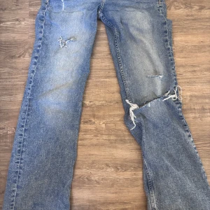 Blå Lågmidjade  slitna jeans  - Säljer ett par blå jeans med raka ben och rejält slitna detaljer på låret och knät. Jeansen har klassisk femficksdesign och är tillverkade i denim. Perfekt för dig som gillar en avslappnad och edgy stil.