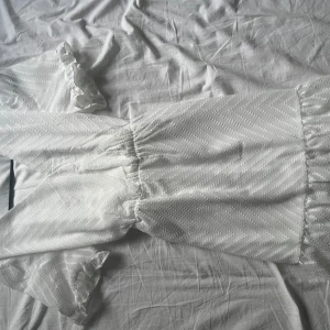 Vit kortklänning  - Superfin vit kortklänning från Vila med subtilt mönster i tyget. Klänningen har v-ringning, korta ärmar med volang och knyt i midjan för en snygg passform. Perfekt för sommarens alla tillfällen och ger en fräsch och luftig känsla.