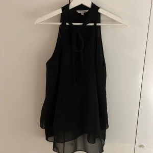Svart ärmlös blus  - säljer en svart blus med justerbar rosett vid halsen och volang nedtill ❣️ tyget är ganska tunt och lite genomskinligt, endast använd en gång
