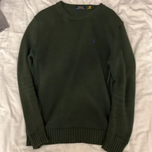 Mörkgrön stickad tröja Polo Ralph Lauren - Mörkgrön stickad tröja från Polo Ralph Lauren med klassisk rund halsringning och diskret blå logga broderad på bröstet. Tröjan har ribbade muddar vid ärmslut och nederkant, perfekt för lager-på-lager under höst och vinter.