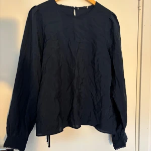 Mörkblå blus med spetsdetaljer - Snygg mörkblå blus med lång ärm och rund halsringning. Blusen har fina spetsdetaljer vid halsen och ärmsluten samt knappar på manschetterna. Tunn och luftig i materialet, perfekt för lager-på-lager. Knytband nertill för extra stilpoäng.