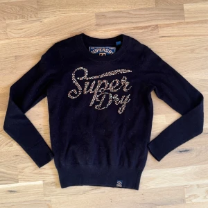 Svart stickad Superdry tröja med stjärnor - Svart stickad tröja från Superdry med lång ärm och rund hals. Framsidan har Superdry-loggan i guldiga och silvriga nitar och stjärnor som ger en snygg och unik look. Perfekt för dig som gillar streetstyle och vill sticka ut lite extra.